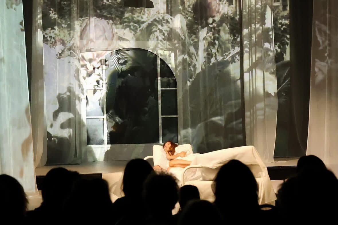 A performer reclining on a white bed beneath projected foliage