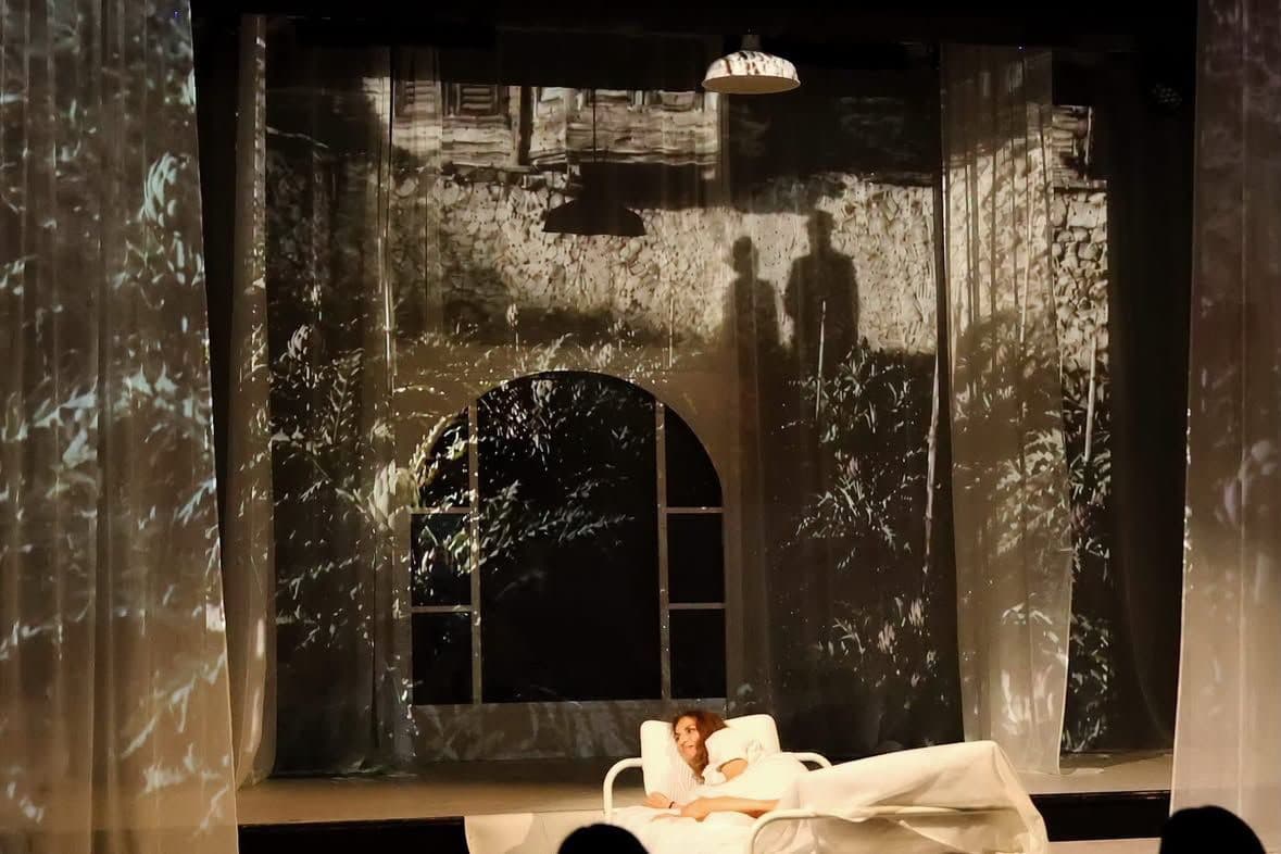 A performer reclining beneath projected foliage and two silhouettes on translucent curtains
