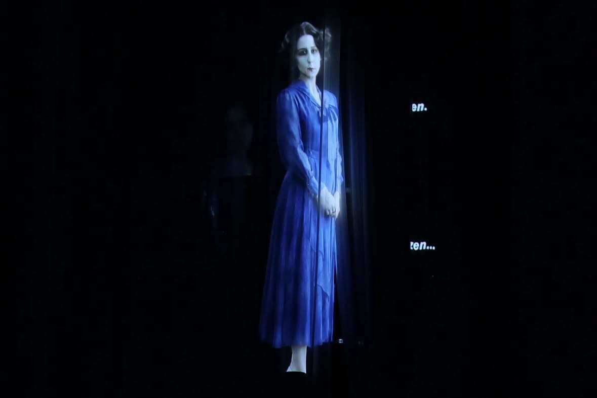 A blue-toned projected portrait of a woman in a dress against a dark stage